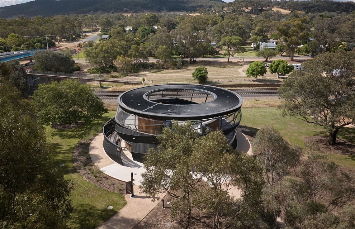 Aerial view of the Ned Kelly Discovery Hub in Glenrowan, Victoria, featuring its circular, modern black structure with an open central skylight, surrounded by trees and pathways, with railway tracks and rural landscape in the background.