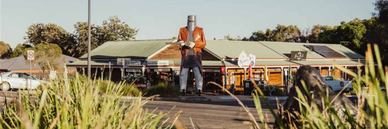 Large Ned Kelly statue in front of a rustic building in Glenrowan, Victoria, with signage and greenery in the foreground.