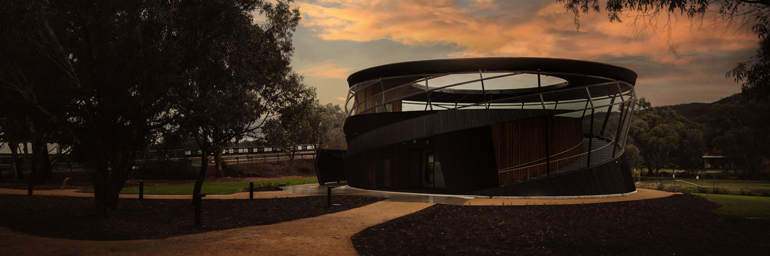 Exterior view of the Ned Kelly Discovery Hub in Glenrowan at sunset, showcasing its circular architectural design surrounded by trees and walking paths.