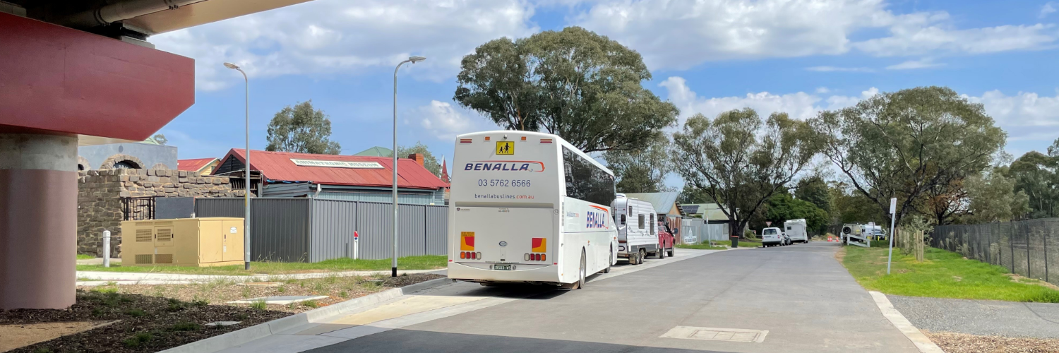 Tour buses parked near the Ned Kelly Discovery Hub in Glenrowan under a bridge, with trees and buildings in the background.